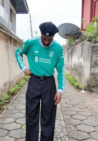 Maillot vert à manches longues avec des accents blancs et bleus, pantalon noir et béret noir. Debout sur un chemin en pierre entre des murs.