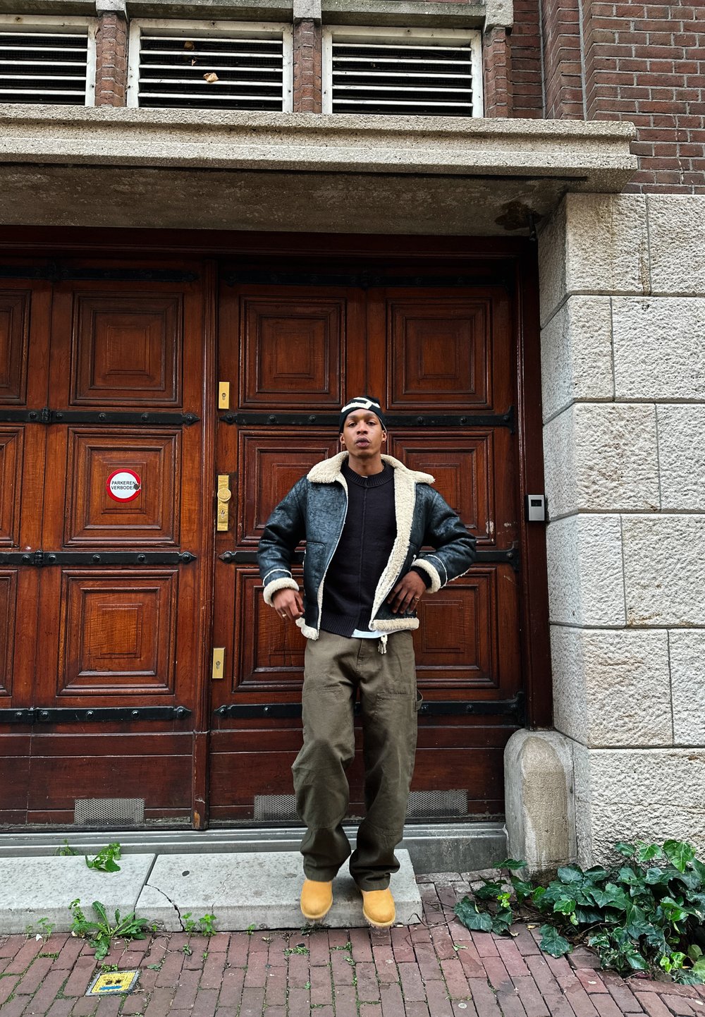 Veste en cuir noir avec col en shearling beige, pull sombre, pantalon cargo olive et bottes jaunes, devant une porte en bois avec des accents en brique.