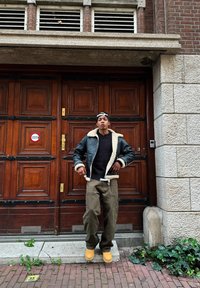 Veste en cuir noir avec col en shearling beige, pull sombre, pantalon cargo olive et bottes jaunes, devant une porte en bois avec des accents en brique.