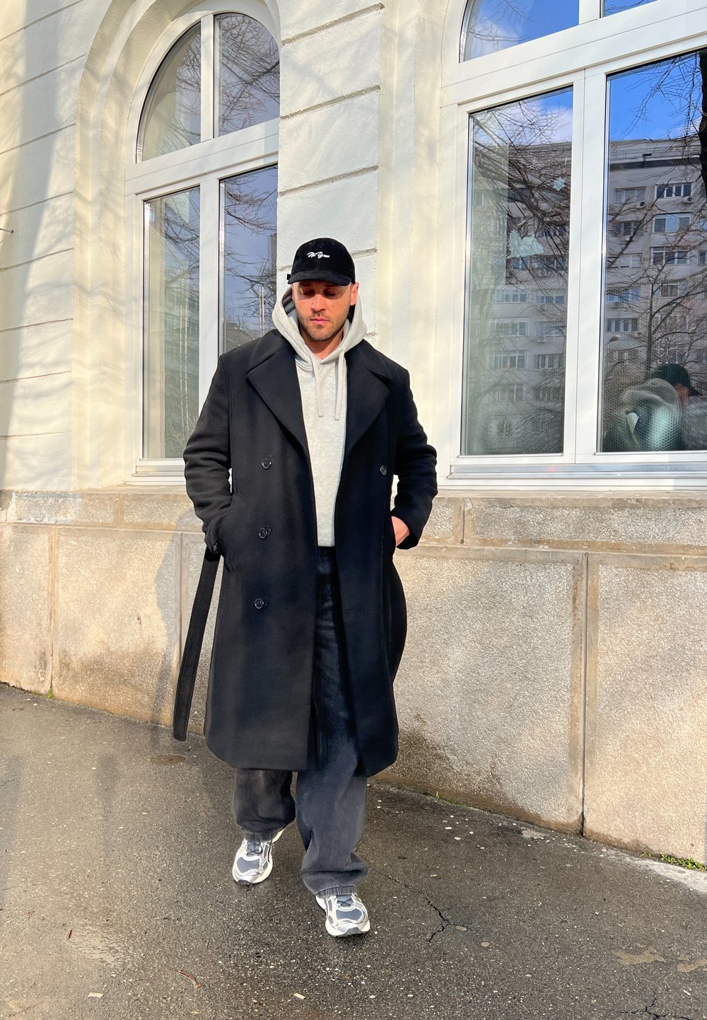 Svart ylleöverrock, grå hoodie, mörka byxor och grå sneakers. Har en dubbelknäppt design och en minimal logotyp på kepsen.