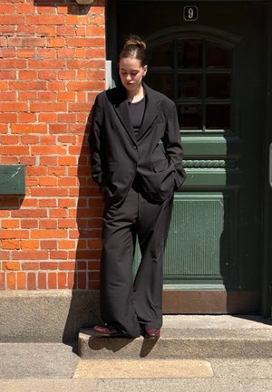 Schwarzer übergroßer Anzug mit Blazer und weiten Hosen, kombiniert mit einem schwarzen Oberteil. Steht an einer Backsteinmauer neben einer grünen Tür.