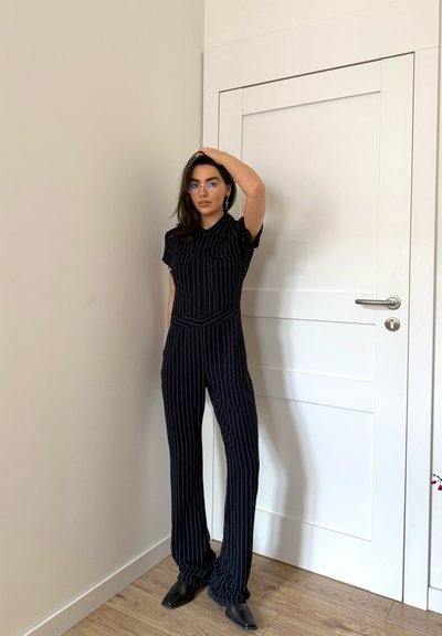 Mujer con gafas, vestida con un mono negro de rayas y zapatos negros puntiagudos, de pie en el interior junto a una puerta blanca con una mano en la cabeza.
