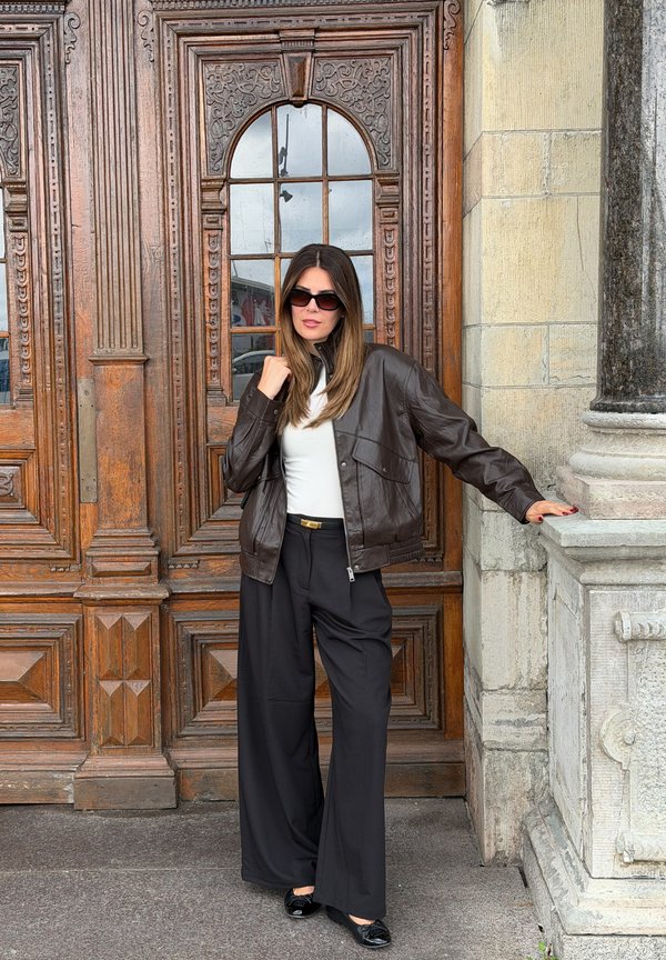 Mujer con gafas de sol, chaqueta de cuero marrón, top blanco y pantalones anchos negros apoyada en un pilar de piedra frente a una puerta de madera ornamentada.