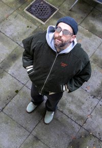 Veste en velours côtelé verte avec un sweat à capuche gris, jean noir et baskets claires. Accessoires : lunettes et bonnet. Debout sur une surface en pierre.