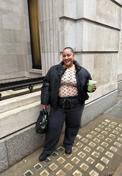 Mujer con chaqueta acolchada negra y top con estampado de luna creciente de pie en la acera sosteniendo una bebida verde y un bolso negro frente a un edificio de piedra.