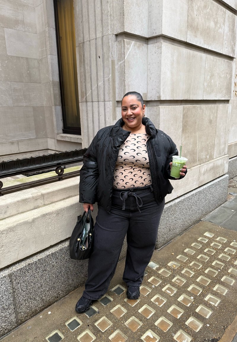 Femme en doudoune noire et haut imprimé d’un croissant de lune, debout sur le trottoir, tenant une boisson verte et un sac à main noir, devant un bâtiment en pierre.