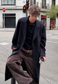 Námornícky modrý vlnený kabát s veľkými klopami, čierny sveter a hnedé široké kordurové nohavice; drží biely šálku kávy. Městské pouličné prostredie.