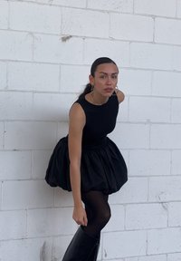 Donna in un vestito nero senza maniche con gonna a sbuffo, collant neri e stivali neri alti posa contro un muro di mattoni bianchi.