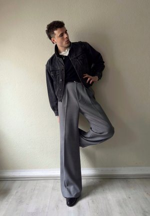 Hombre apoyado contra una pared beige, vistiendo una chaqueta de mezclilla negra, camisa negra, pantalones gris de pierna ancha y zapatos negros, con una pierna doblada.