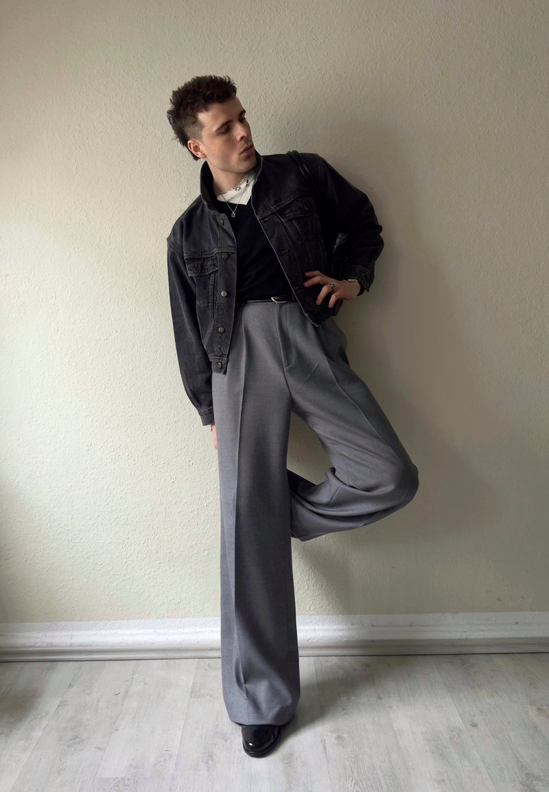 Mann, der an eine beige Wand lehnt, trägt schwarze Jeansjacke, schwarzes Hemd, graue weite Hose und schwarze Schuhe, mit einem angewinkelten Bein.