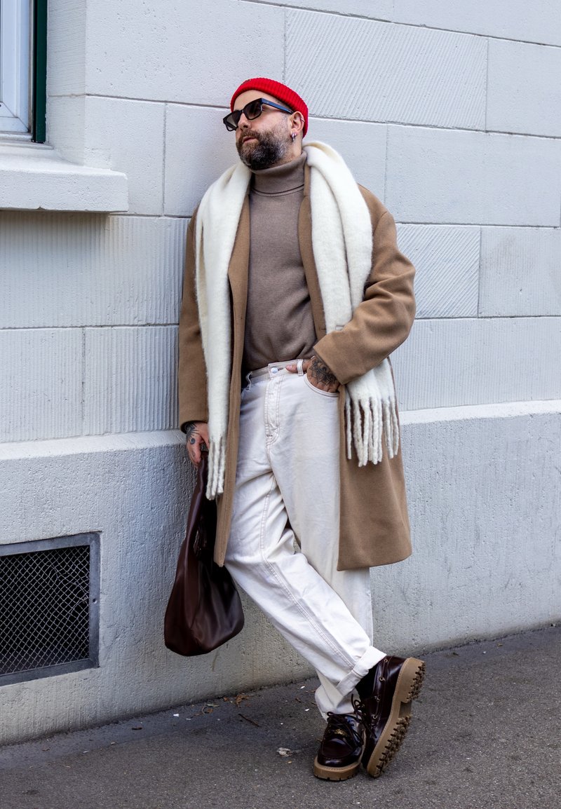 Manteau en laine marron, col roulé beige, écharpe crème, pantalon blanc, sac en cuir marron, chaussures foncées à semelles épaisses, bonnet rouge, lunettes de soleil.