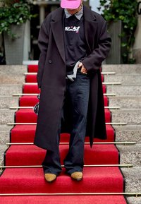 Melna garā jaka virs melna sporta krekla ar baltu uzrakstu, kombinēta ar ziliem džinsiem un brūnām kurpēm, stāvot uz sarkanām paklāja kāpnēm.