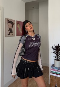 Jauna persona ar sarkaniem matiem, tērpusies melnā, izgaismotā mini svārkā, slāņota sporta augšdaļā, ar galvas lentu un melnu, uzadamētu plecu somu, iekštelpās pie sienas mākslas.