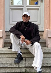 Blazer nero sopra un dolcevita nero, pantaloni bianchi e scarpe nere massicce. Accessori includono anelli e una collana d'argento. Sfondo: porta bianca.