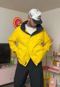 Giacca impermeabile gialla con colletto blu, tasche frontali e chiusura con zip. Pantaloni neri e un cappellino bianco con un logo.