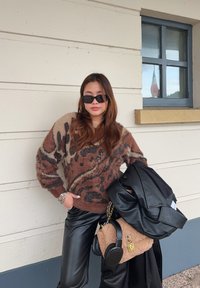 Καφέ και μπεζ πουλόβερ με ζωντανό σχέδιο ζώου, μαύρα δερμάτινα παντελόνια και μια μαύρη τσάντα ώμου. Το πουλόβερ έχει χαλαρή εφαρμογή και υφή.