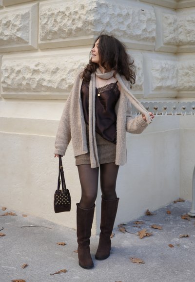 Cárdigan beige de punto sobre un camisón de satén oscuro, combinado con una minifalda marrón texturizada. Botas marrones hasta la rodilla y un bolso pequeño con detalles dorados.
