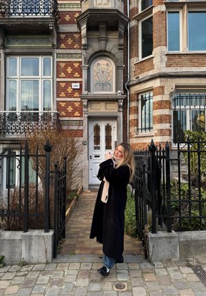 Mujer con abrigo negro y bufanda beige de pie en una acera de adoquines, ajustándose el cabello frente a una casa adosada de ladrillo ornamentado con reja de hierro.