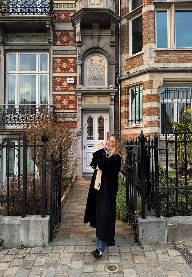 Femme en manteau noir et écharpe beige debout sur un trottoir pavé, ajustant ses cheveux devant une maison de ville en brique ornée avec une grille en fer.