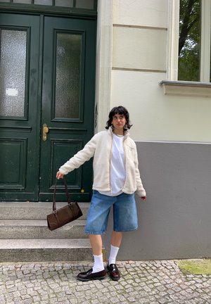 Chaqueta de forro polar blanca con cremallera, camiseta blanca, pantalones cortos de mezclilla, bolso marrón, calcetines blancos, zapatos negros. De pie junto a puertas verdes en escaleras de piedra.