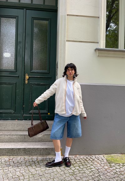 Chaqueta de forro polar blanca con cremallera, camiseta blanca, pantalones cortos de mezclilla, bolso marrón, calcetines blancos, zapatos negros. De pie junto a puertas verdes en escaleras de piedra.