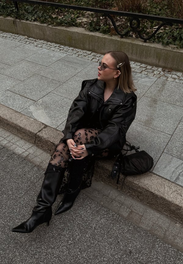 Femme portant une veste en cuir noire, des collants transparents à motifs, des bottes noires et des lunettes de soleil, assise sur un trottoir avec un sac noir à côté d'elle.