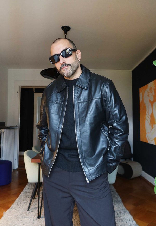 Giacca nera in pelle con chiusura a zip, colletto e due tasche laterali. Indossata sopra un maglione nero e abbinata a pantaloni scuri.