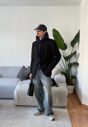 Hombre con blazer negro de rayas, sudadera con capucha azul marino, vaqueros holgados, zapatillas, gorra y gafas de sol, de pie en la sala de estar sosteniendo una bolsa negra.
