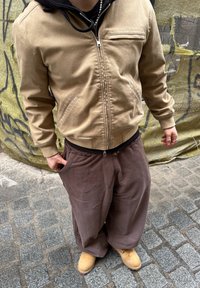 Jaqueta bomber bege com um bolso frontal, combinada com calças largas castanhas e botas amarelas claras. Superfície texturizada e ajuste descontraído.