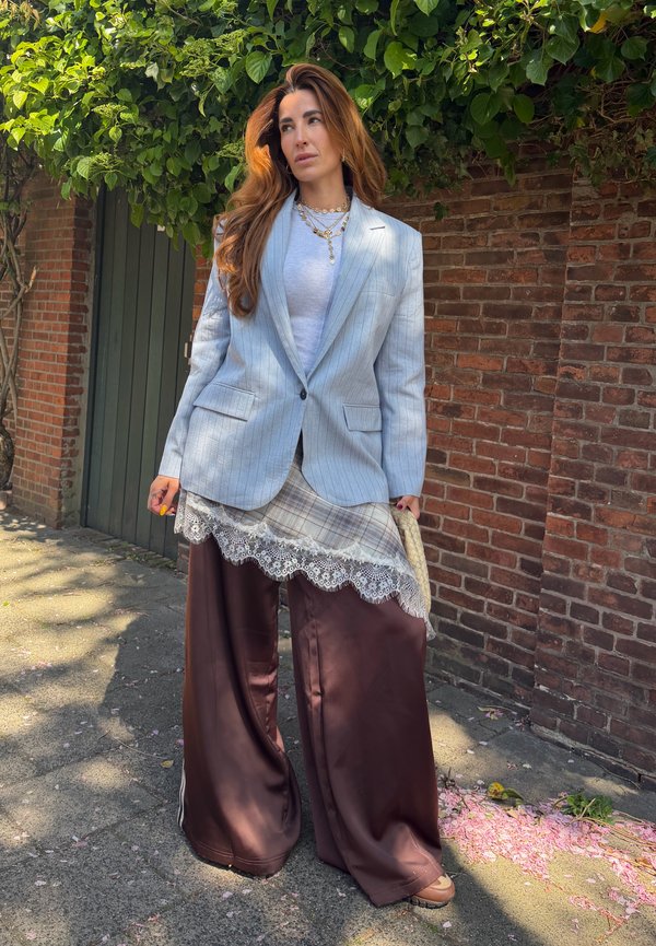 Frau mit langen braunen Haaren, die ein geschichtetes Outfit trägt: hellblaues Nadelstreifen-Blazer, weißes Oberteil, karierter Rock mit Spitzenbesatz und weite braune Hosen im Freien.