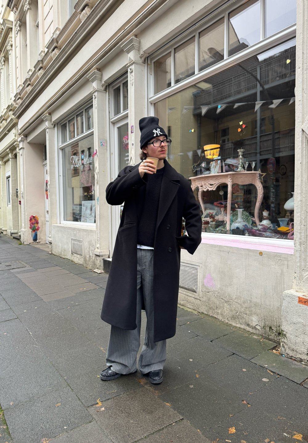 Cappotto di lana nero, maglione nero a maglia, pantaloni a gamba larga grigi a righe, scarpe nere; tiene in mano un bicchiere di carta; in piedi su un marciapiede bagnato vicino a una vetrina.