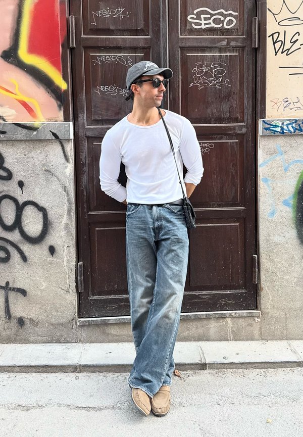 Hombre con camisa blanca de manga larga, vaqueros azules y gorra gris apoyado contra una pared cubierta de grafitis con una puerta de madera oscura detrás de él.