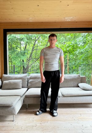 Camisa gris de mangas cortas, pantalones sueltos negros y sandalias negras con acentos plateados. Sofá en tonos neutros con suelo de madera clara de fondo.