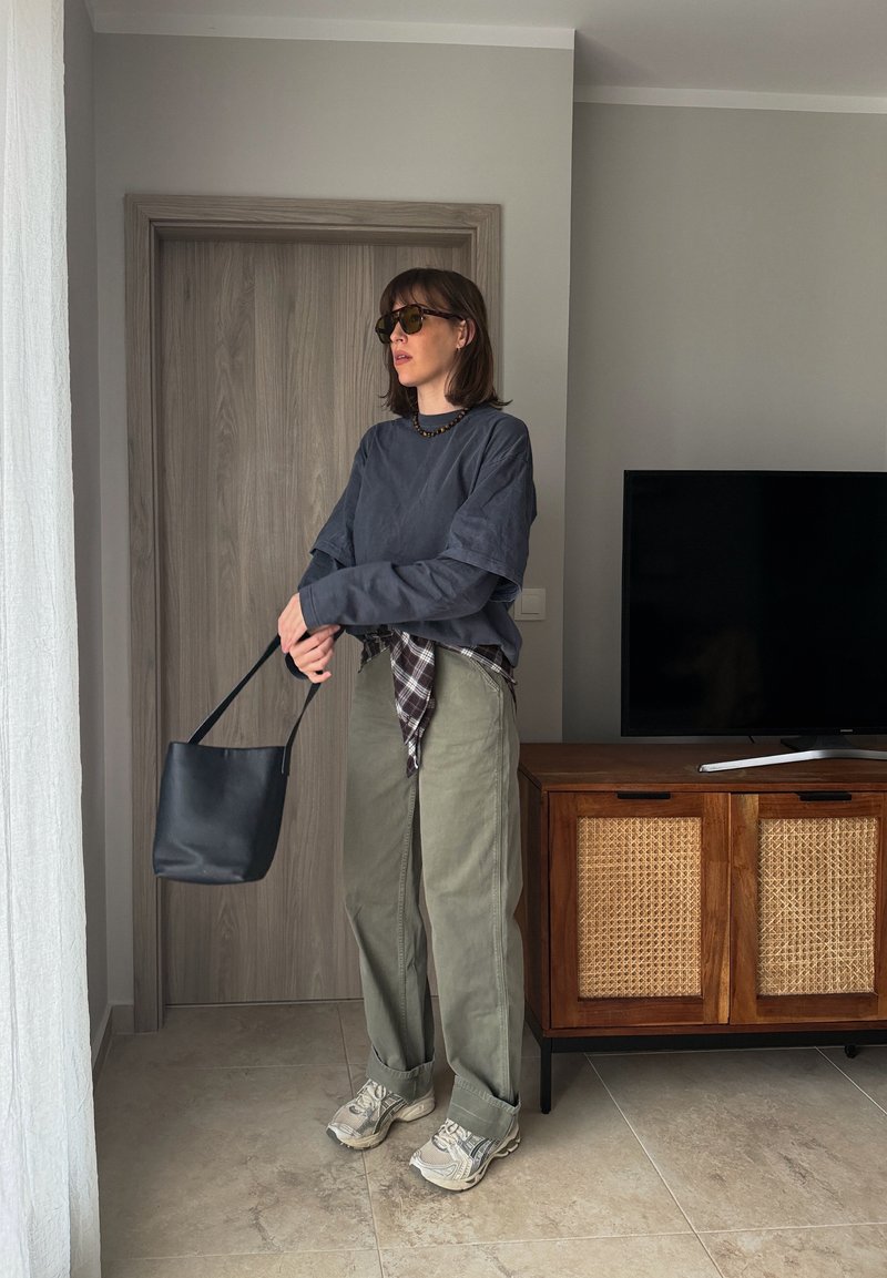 Moteris su akiniais nuo saulės, pilka palaidine, žaliai kelnėmis ir sportbačiais, laikanti juodą rankinę svetainėje prie medinių durų ir TV spintelės.