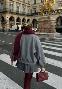 Szary, oversize sweter, bordowa chusta z frędzlami, plisowana szara spódnica, bordowe rajstopy i fakturowana bordowa torebka. Miejska sceneria z pomnikiem.
