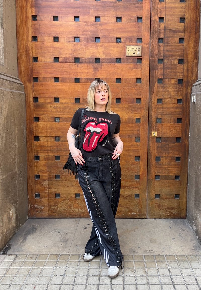 Fekete Rolling Stones pólót és fekete fűzős nadrágot viselő nő áll egy nagy, négyszögletes kivágásokkal díszített faajtó előtt.