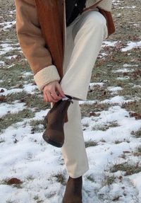 Personne à l'extérieur dans la neige ajustant une botte marron, portant un manteau en peau retournée beige et un pantalon blanc cassé, debout sur de l'herbe partiellement couverte de neige.