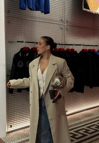 Beige dobbeltknappet frakke over hvid bluse, blå jeans, holder en metallisk clutch og en kaffekop, med sorte jakker hængende i baggrunden.