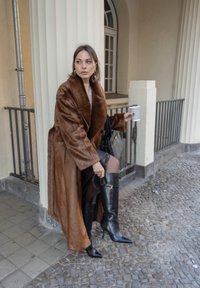 Long manteau en fausse fourrure marron, bottes noires en cuir montantes jusqu'aux genoux avec des détails dorés, sac à main noir, tenant une tasse de café, sur un fond en pierre.