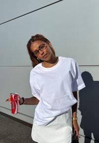 T-shirt bianca oversize con scollo rotondo, abbinata a una mini gonna bianca. La persona tiene in mano una lattina di soda rossa. Lo sfondo è grigio.