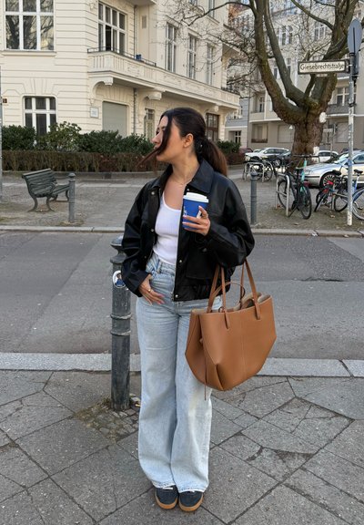 Chaqueta de cuero negra, camiseta sin mangas blanca, jeans anchos de color azul claro, bolso de cuero marrón, sosteniendo una taza de café azul, de pie en una acera.