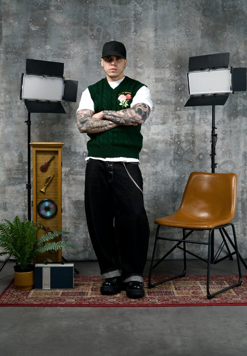 Junger Mann mit tätowierten Armen verschränkt, trägt eine grüne Weste und eine schwarze Kappe, steht zwischen Studiolichtern, einem Stuhl und vintage Musikdekoration.