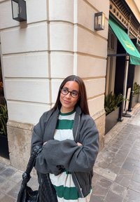 Femme aux longs cheveux bruns et lunettes, portant une chemise rayée et une veste grise, se tient à un coin de rue carrelé près d'un bâtiment avec des plantes.