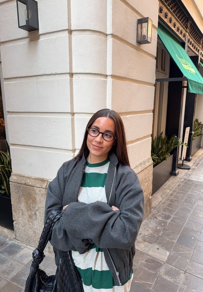 Mujer con cabello largo y castaño y gafas, vestida con una camisa de rayas y una chaqueta gris, de pie en una esquina de calle con baldosas cerca de un edificio con plantas.