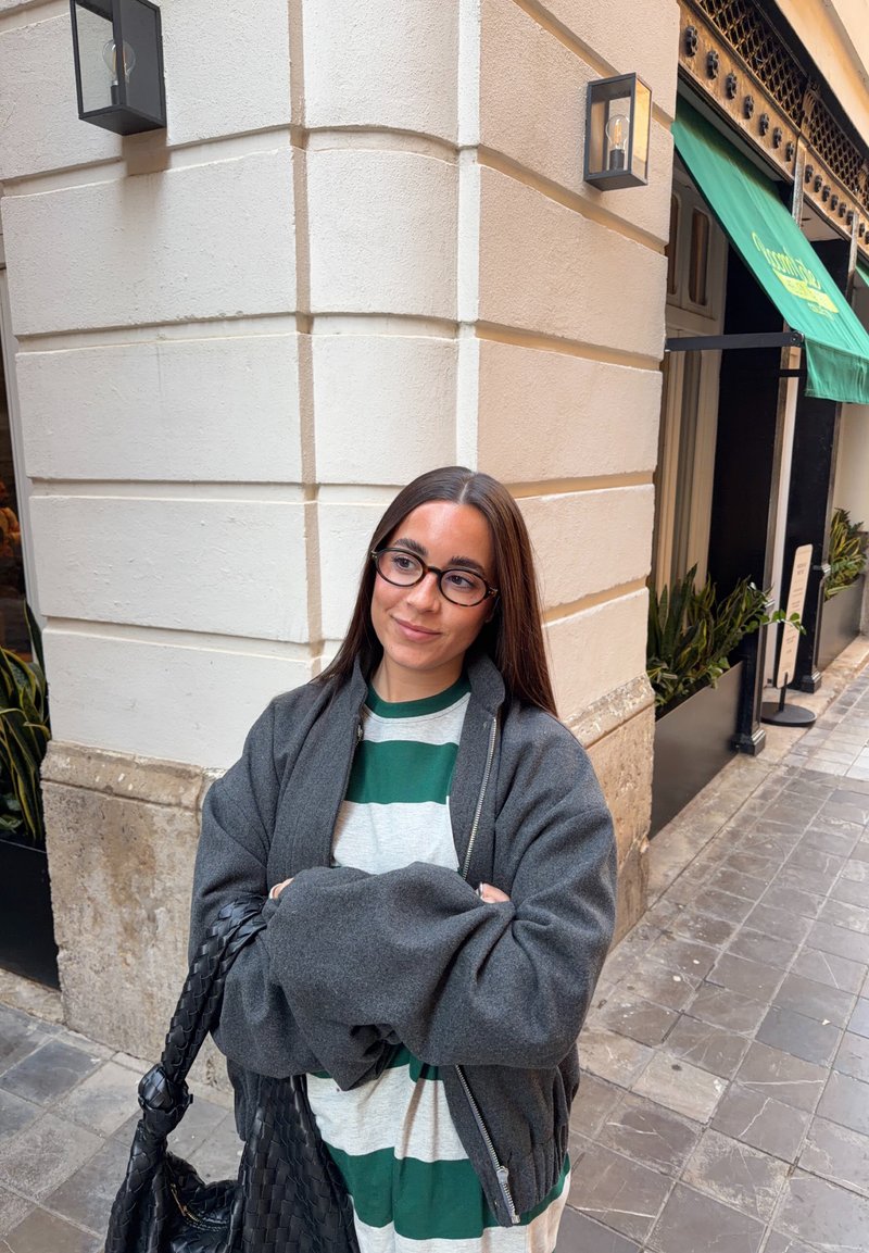 Femme aux longs cheveux bruns et lunettes, portant une chemise rayée et une veste grise, se tient à un coin de rue carrelé près d'un bâtiment avec des plantes.