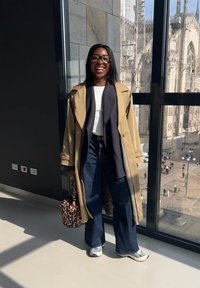 Tan trenchcoat över en marinblå cardigan och vit topp, vida denimjeans och handväska med djurmönster. Sneakers med texturerad detalj.