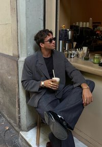 Sivina oversized blazer in sivi in barvni hlače, črna majica, sončna očala, drži pijačo, sedi na marmorju s stolom z zlatimi akcenti.