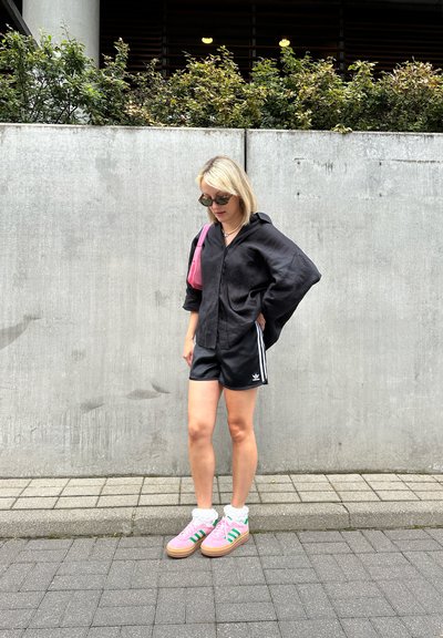 Camisa negra de corte holgado con botones, pantalones cortos negros con rayas blancas, zapatillas rosas con rayas verdes y calcetas blancas. Fondo de concreto.