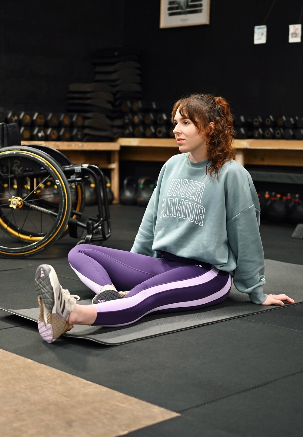 Athlète en sweat-shirt bleu clair et legging violet avec des bandes blanches, assise sur un tapis gris à côté d'un fauteuil roulant noir et jaune dans une salle de sport.