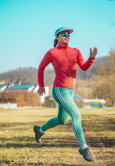 Chaqueta roja con cremallera, leggins verdes con rayas blancas, gafas de sol y una gorra; corriendo por un sendero de hierba bajo un cielo azul claro.